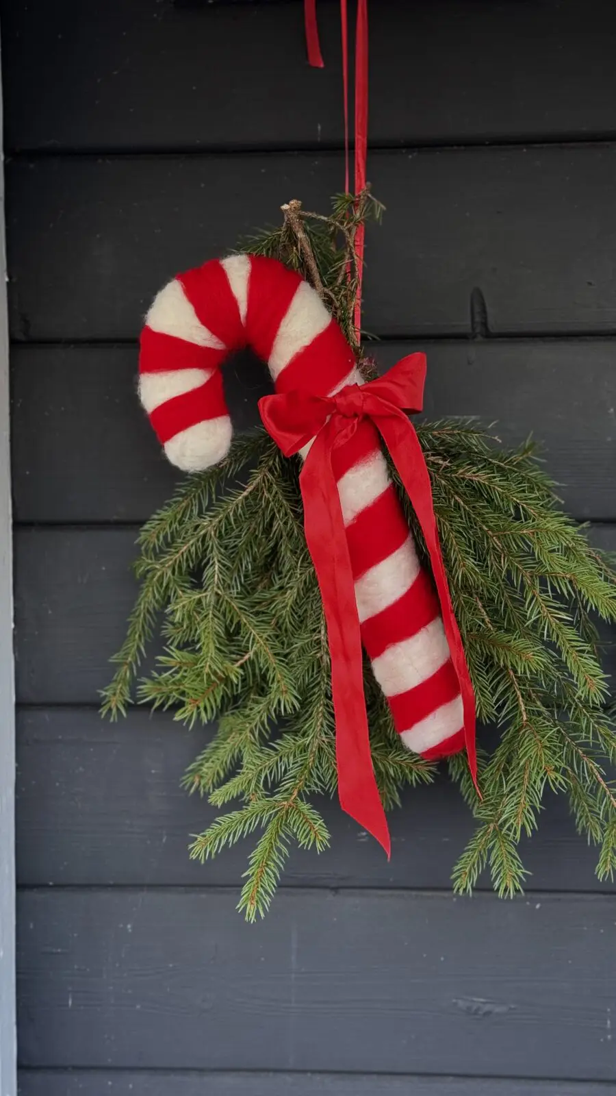 DIY julkrans av granris med stor nålfiltad polkagris och röd rosett hängandes på svart ytterdörr