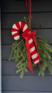 DIY julkrans av granris med stor nålfiltad polkagris och röd rosett hängandes på svart ytterdörr