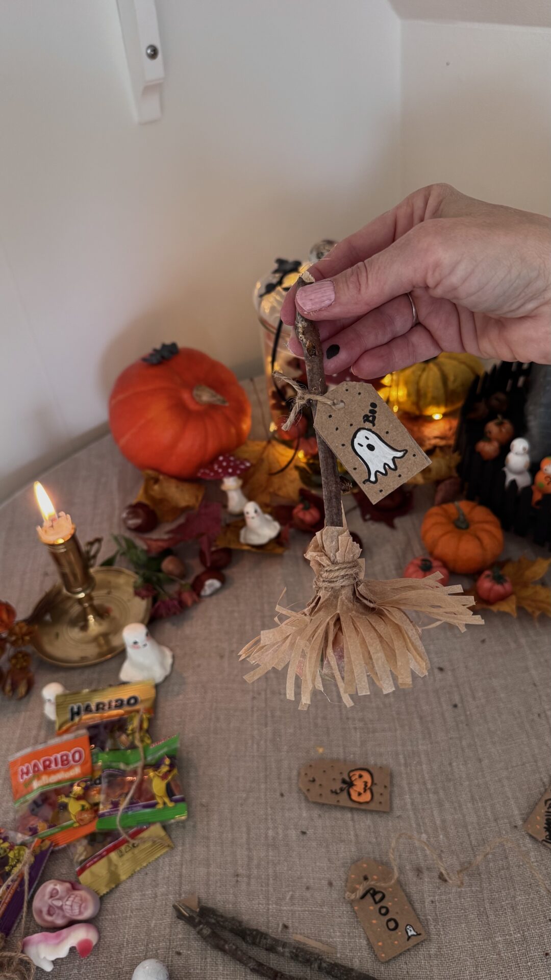 Hand som håller upp en färdig häxkvast av pinne och bakplåtspapper, dekorerad med etikett och snöre, i höstlig Halloweenmiljö med pumpor och ljus.
